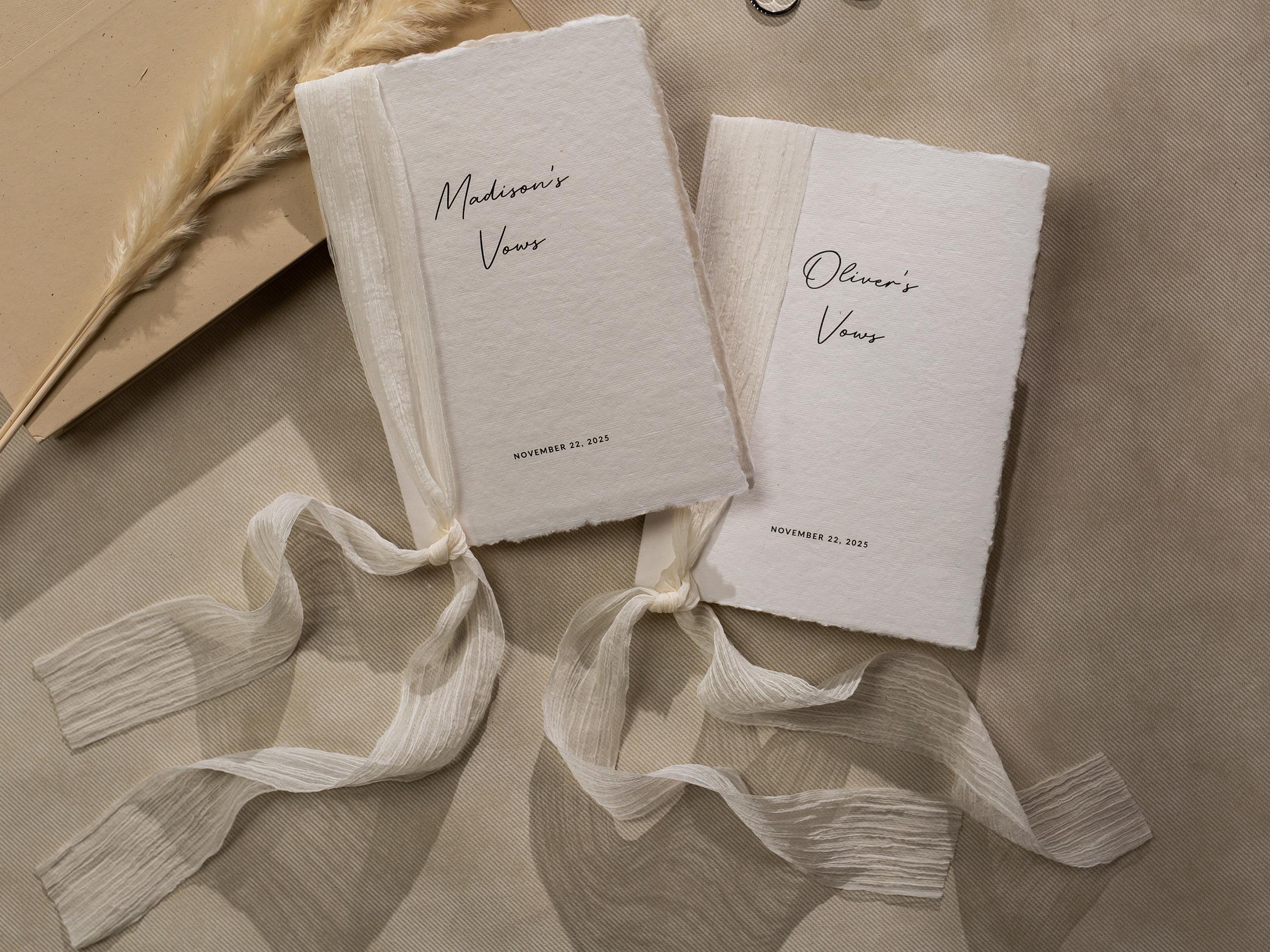 May include: Two white paper vow books with a cream colored ribbon tied around them. The books are labeled &#39;Madison&#39;s Vows&#39; and &#39;Oliver&#39;s Vows&#39; with the date &#39;November 22, 2023&#39; printed below the names. The books are on a beige surface.