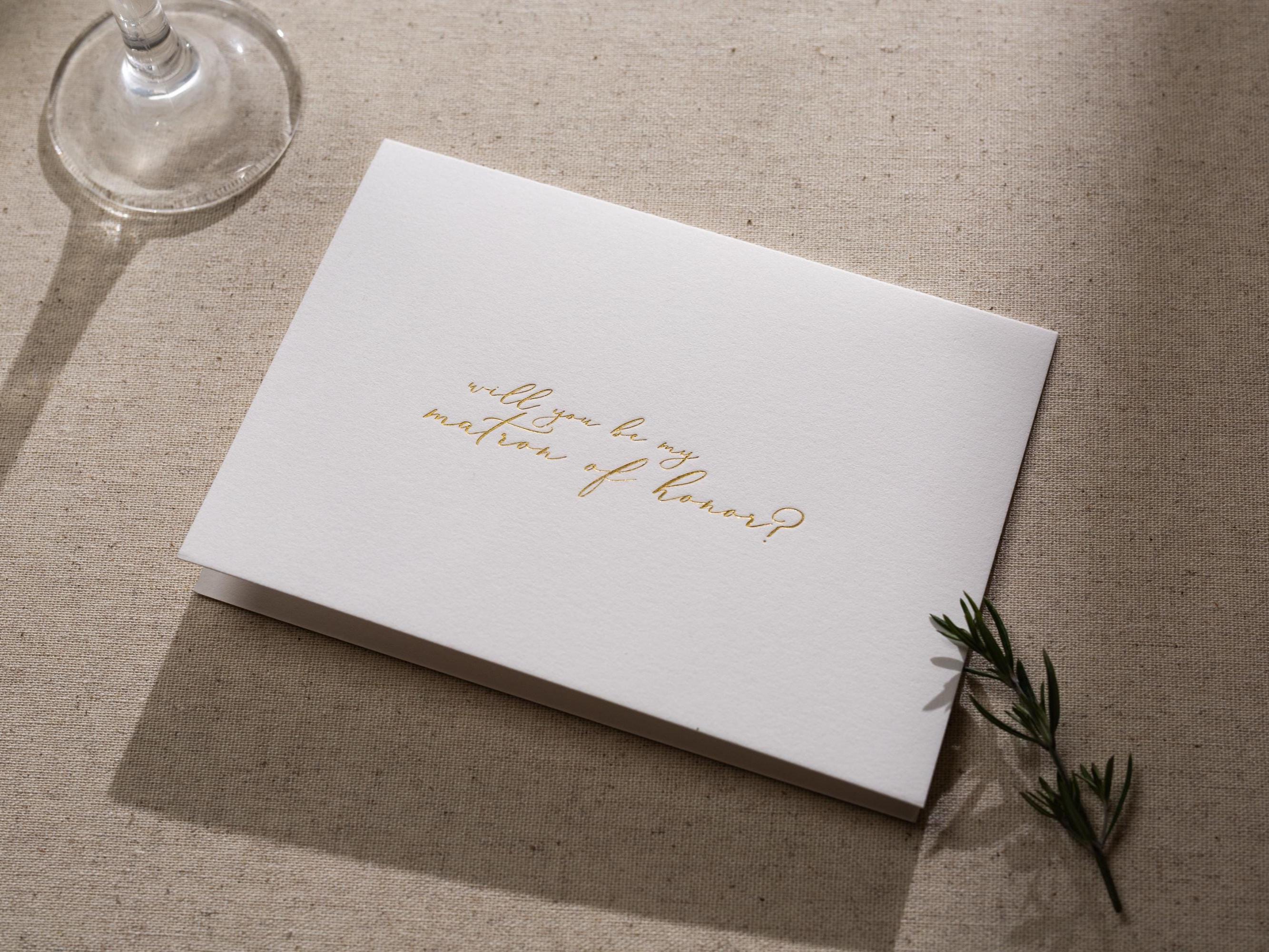 May include: A white card with gold lettering that reads &#39;Will you be my matron of honor?&#39;