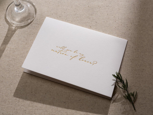 May include: A white card with gold lettering that reads 'Will you be my matron of honor?'