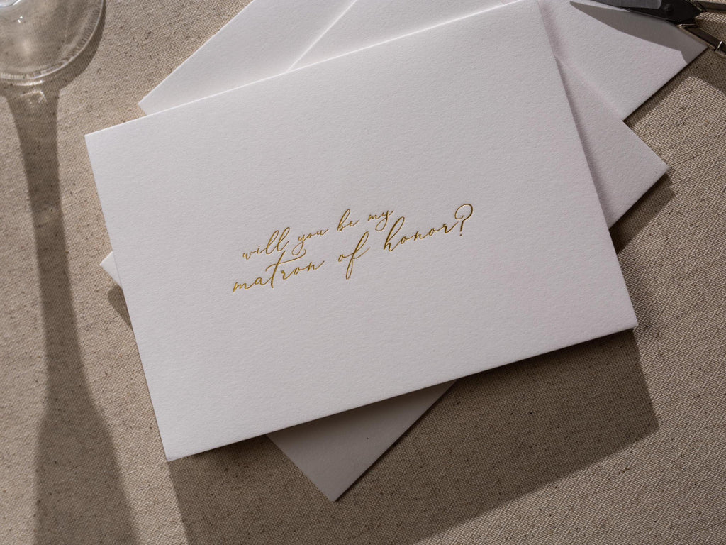 May include: A white card with gold lettering that says &#39;Will you be my matron of honor?&#39;