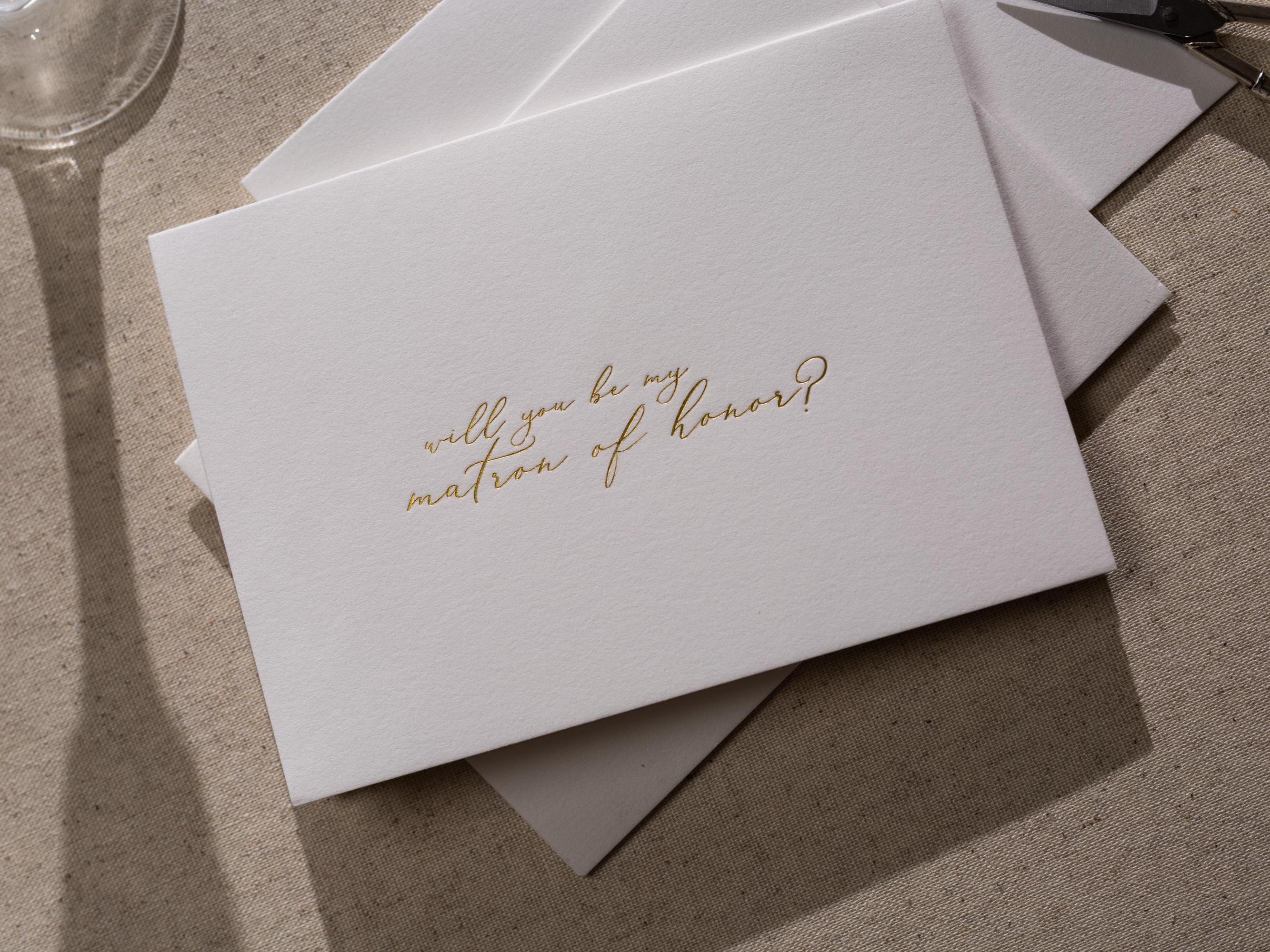 May include: A white card with gold lettering that says &#39;Will you be my matron of honor?&#39;