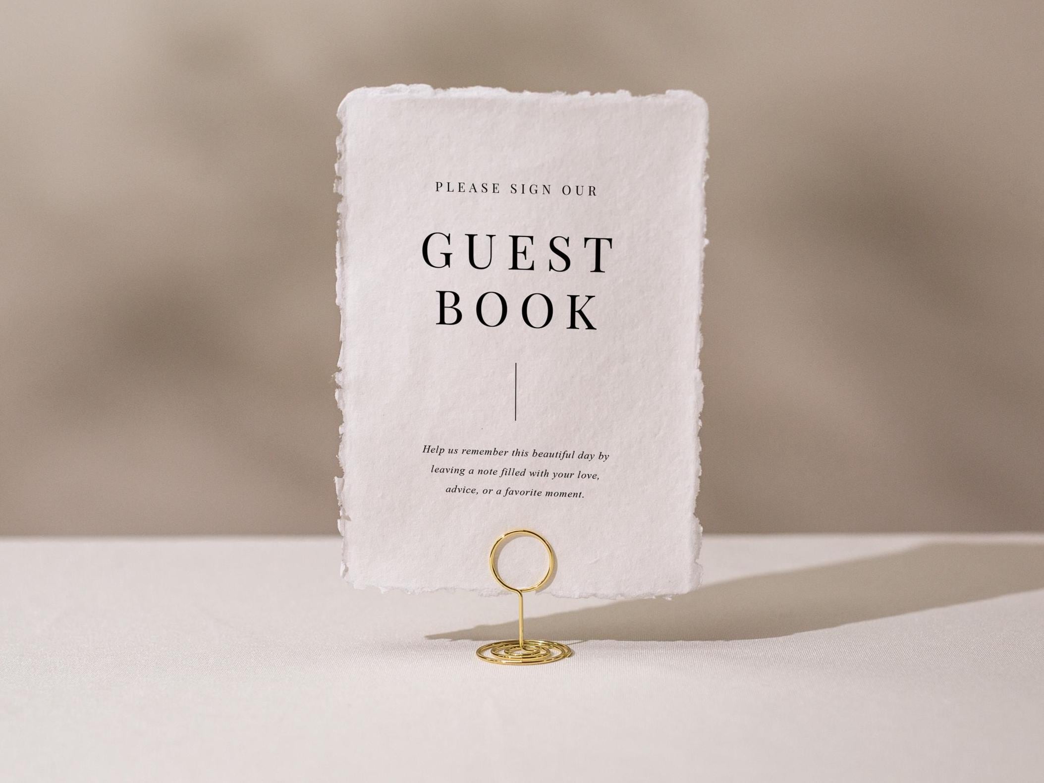 May include: A rectangular guest book sign with a torn edge design. The sign is white with black text that reads 'PLEASE SIGN OUR GUEST BOOK'. Below the text is a vertical line. The sign is held by a gold metal holder. The sign includes text that reads 'Help us remember this beautiful day by leaving a note filled with your love, advice, or a favorite moment.'