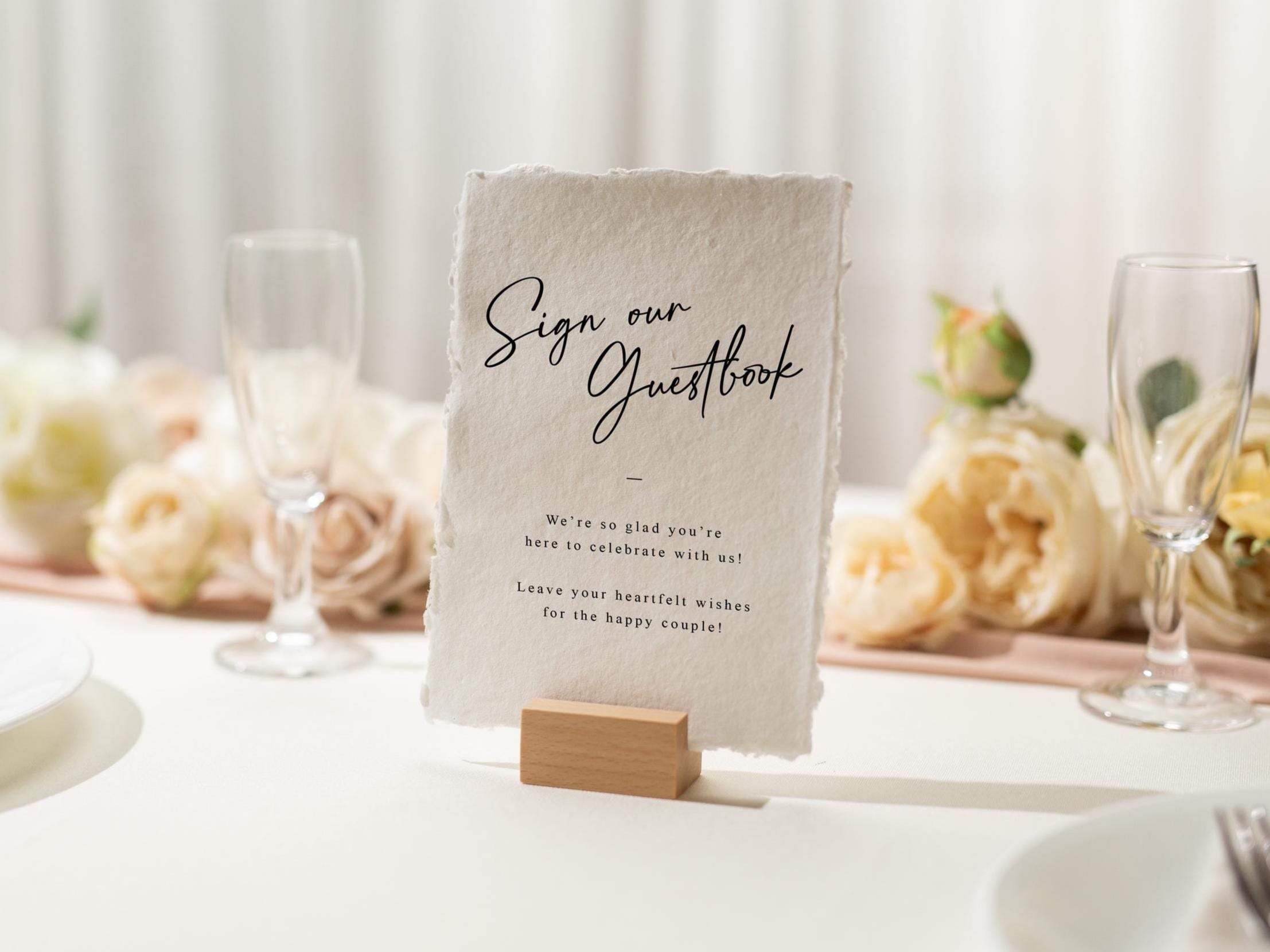 May include: A wedding guestbook sign on a white table. The sign is a textured, off-white paper with the words "Sign our Guestbook" in elegant script. The sign is held by a small wooden block. Champagne flutes and floral arrangements are in the background.