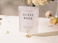 May include: A white textured paper guest book sign with the words "GUEST BOOK" in black. The sign is held by a gold wire holder. The sign also includes the text "Please sign our" and a short message below the title.