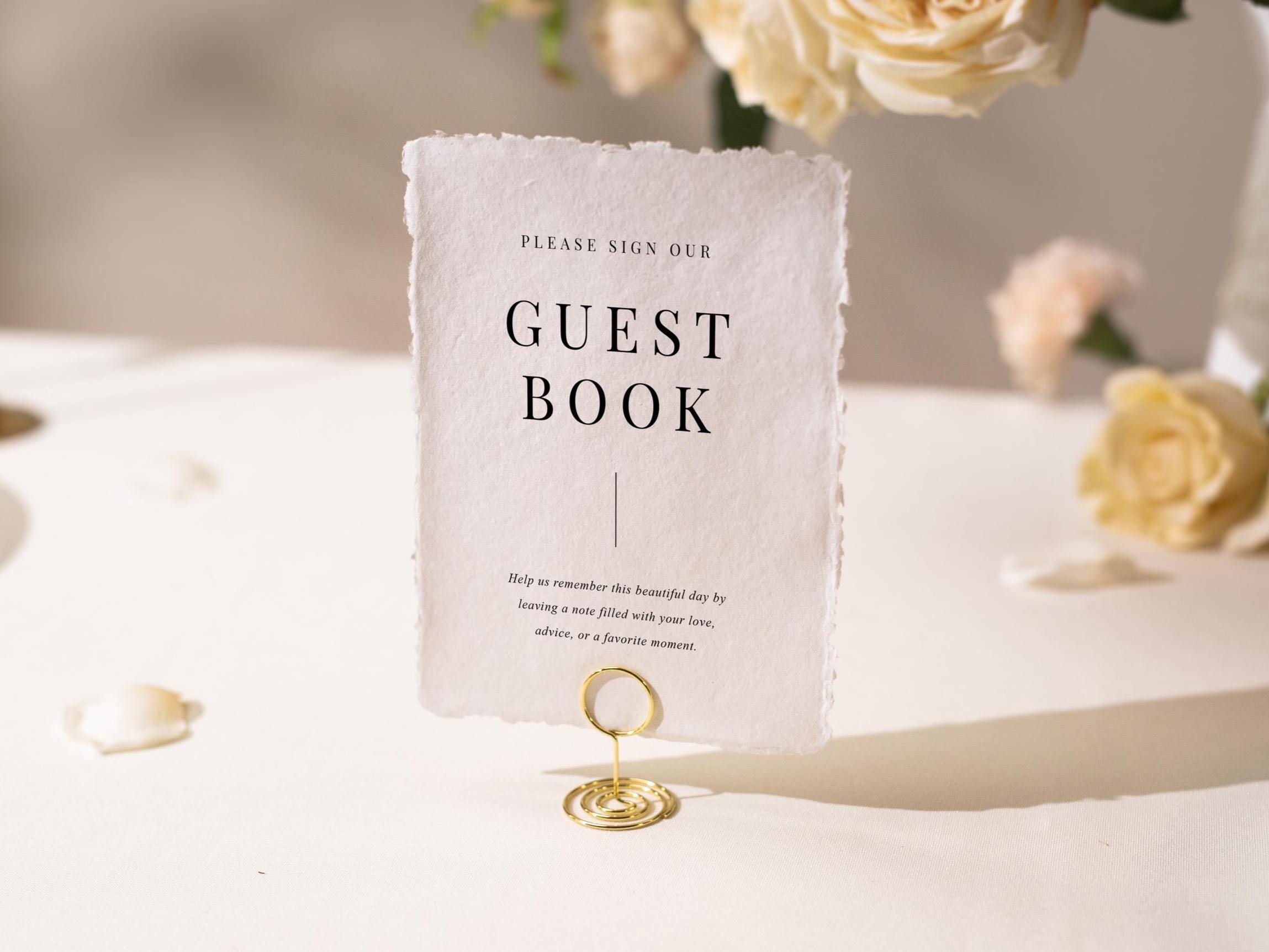 May include: A white textured paper guest book sign with the words "GUEST BOOK" in black. The sign is held by a gold wire holder. The sign also includes the text "Please sign our" and a short message below the title.