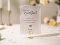 May include: A rectangular, off-white sign with the words "PHOTO guestbook" in elegant script. The sign includes instructions and a camera icon, held by a gold wire holder. The sign is for a wedding or event.