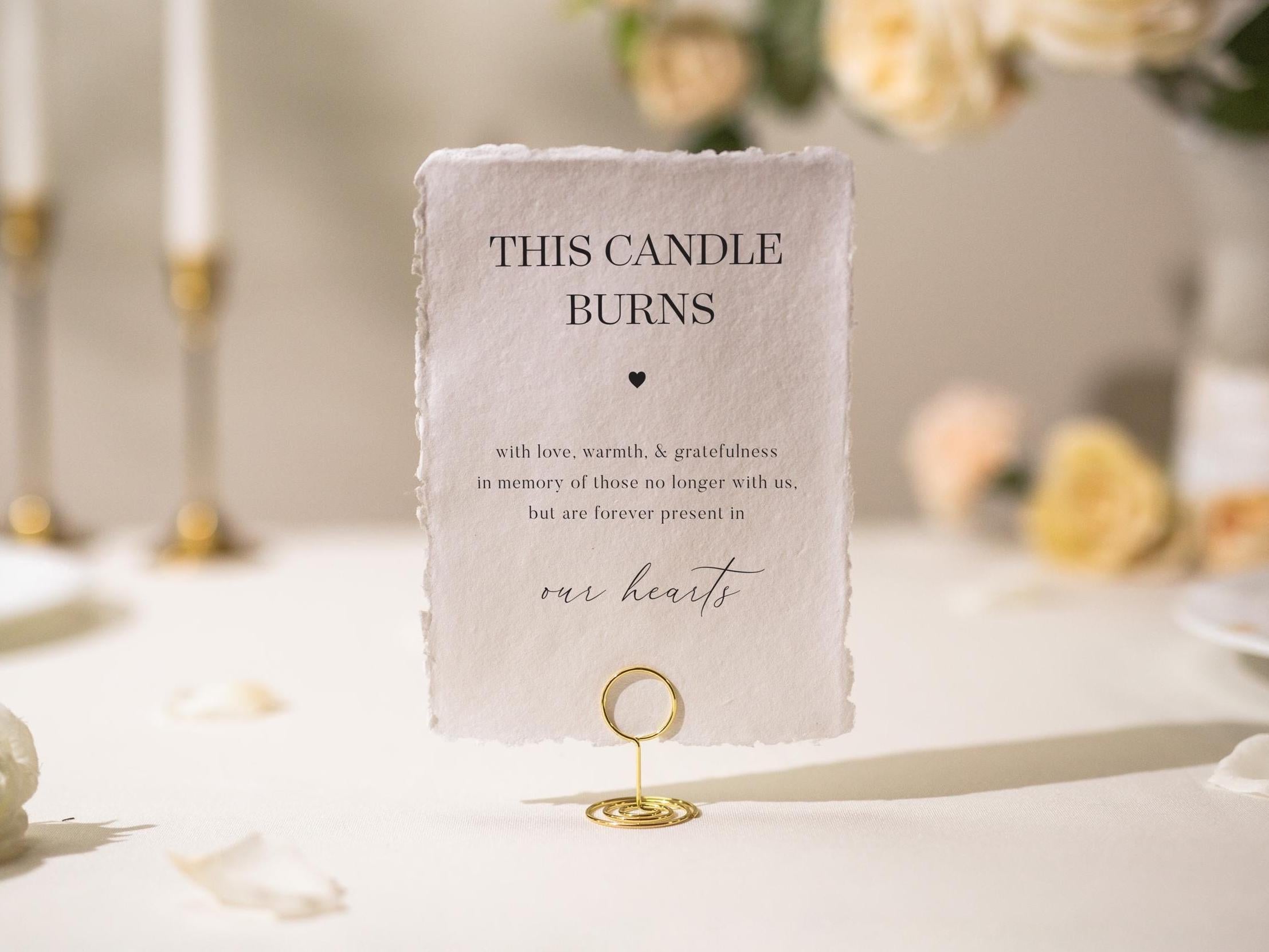 May include: A white paper sign with the text "THIS CANDLE BURNS" and a heart symbol. The sign is held by a gold wire holder. The text below reads "with love, warmth, & gratefulness in memory of those no longer with us, but are forever present in our hearts."