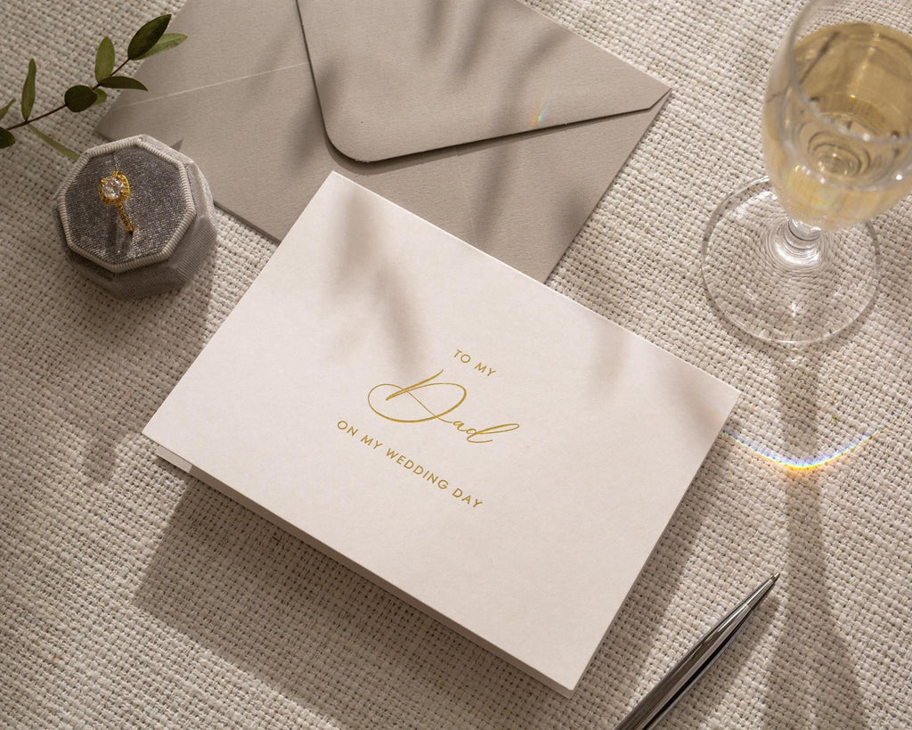 May include: A wedding card with the message "To My Dad On My Wedding Day" in gold script. The card is on a textured surface, with a gray envelope, a ring box, a pen, and a glass of champagne.