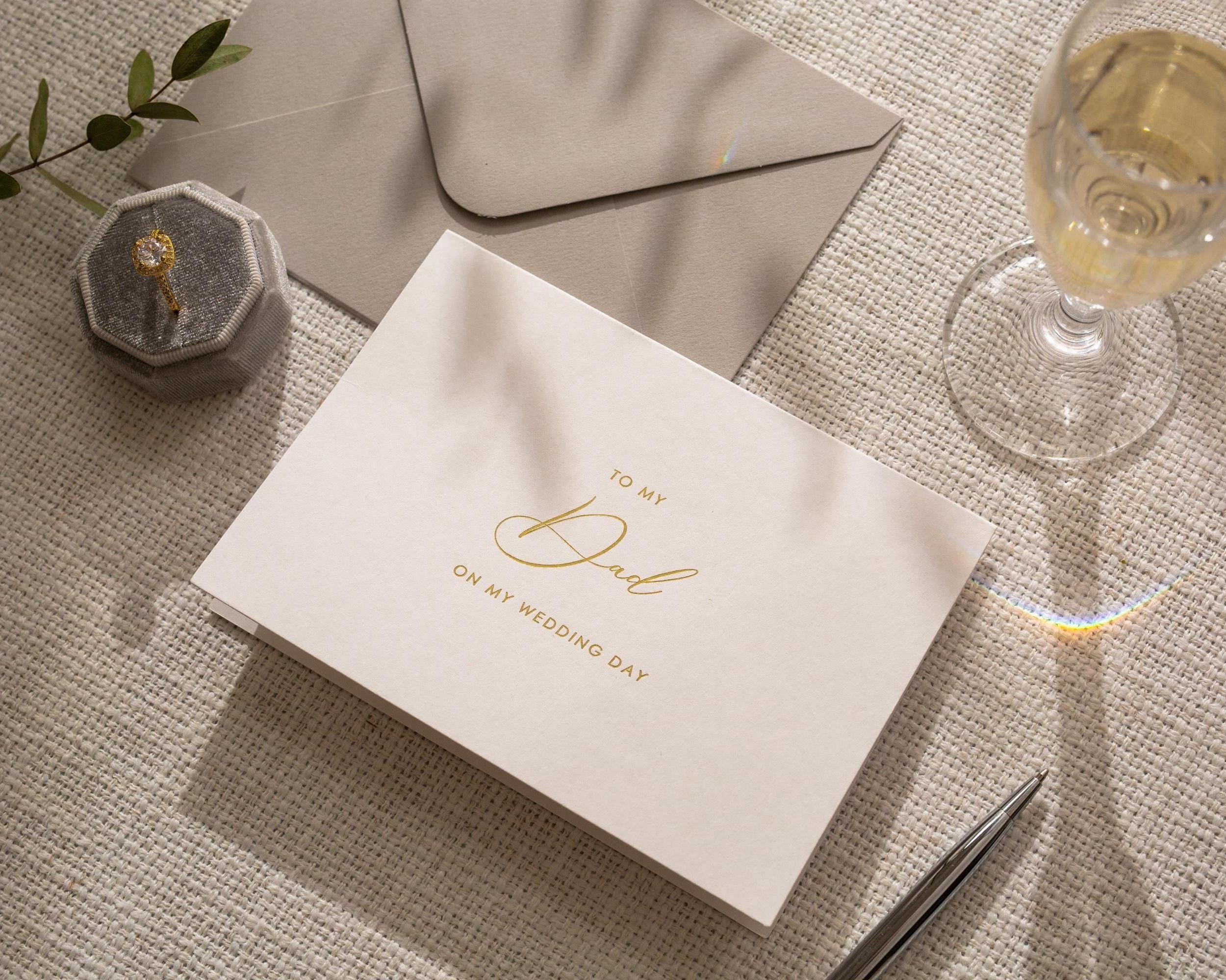 May include: A wedding card with the message "To My Dad On My Wedding Day" in gold script. The card is on a textured surface, with a gray envelope, a ring box, a pen, and a glass of champagne.