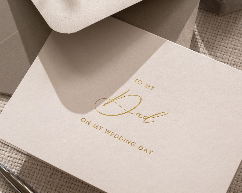 May include: A cream-colored greeting card with gold script that reads "TO MY Dad ON MY WEDDING DAY." The card is partially visible, with a matching envelope in the background. The card is simple and elegant.
