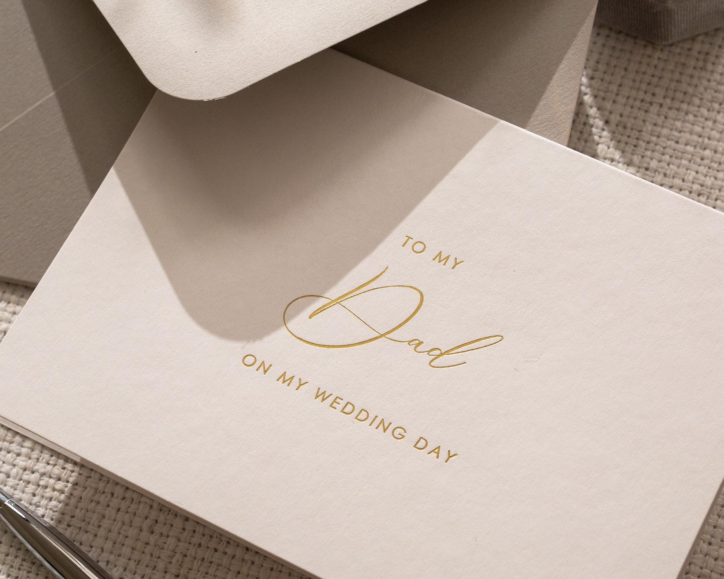 May include: A cream-colored greeting card with gold script that reads "TO MY Dad ON MY WEDDING DAY." The card is partially visible, with a matching envelope in the background. The card is simple and elegant.