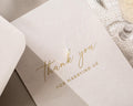 May include: A close-up of a light pink wedding thank you card with gold foil lettering. The card reads "thank you" in elegant script, with "FOR MARRYING US" below in a smaller, sans-serif font.