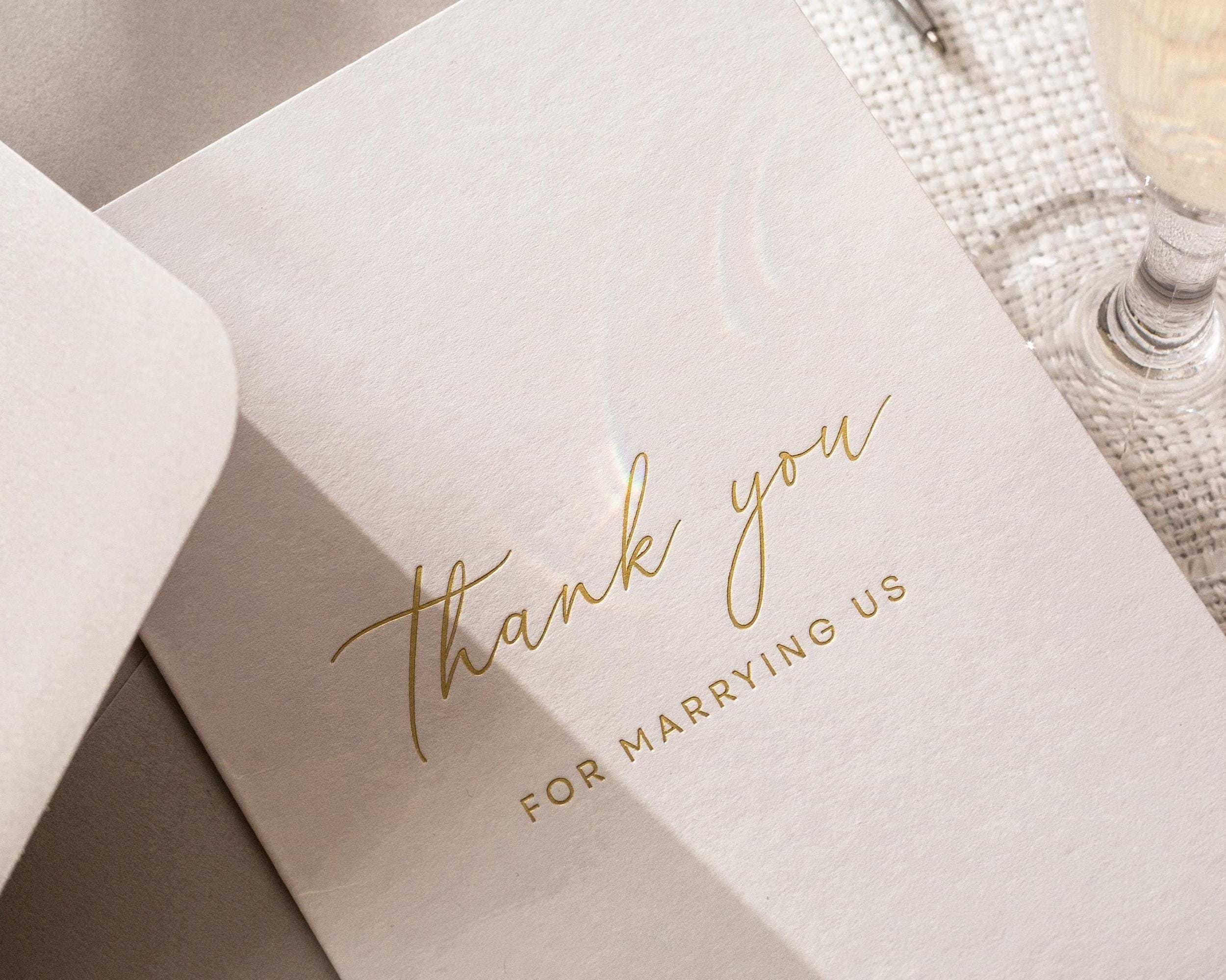 May include: A close-up of a light pink wedding thank you card with gold foil lettering. The card reads "thank you" in elegant script, with "FOR MARRYING US" below in a smaller, sans-serif font.