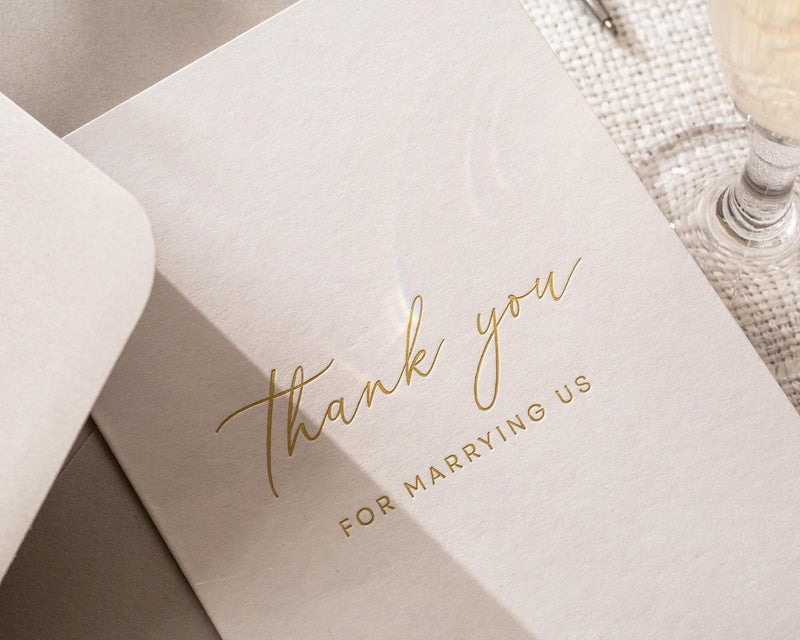 May include: A close-up of a light pink wedding thank you card with gold foil lettering. The card reads "thank you" in elegant script, with "FOR MARRYING US" below in a smaller, sans-serif font.