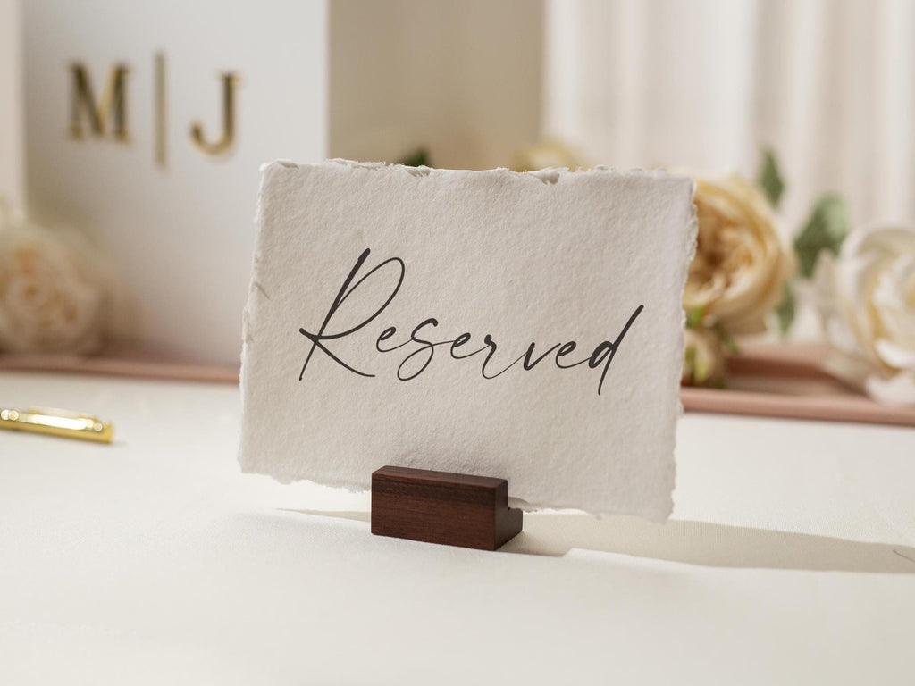 May include: A white card with the word "Reserved" in elegant black script, held by a small, dark wooden block. The card has a textured, deckled edge. The background includes a white box with gold initials and flowers.
