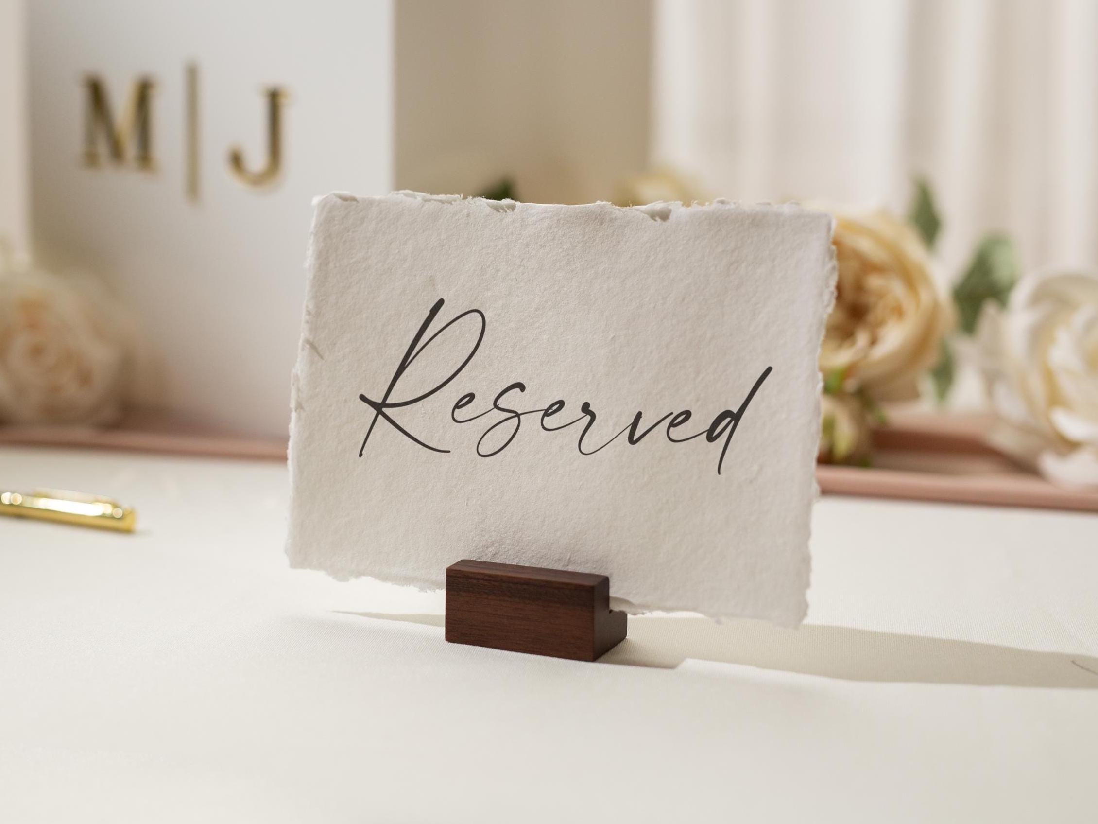 May include: A white card with the word "Reserved" in elegant black script, held by a small, dark wooden block. The card has a textured, deckled edge. The background includes a white box with gold initials and flowers.