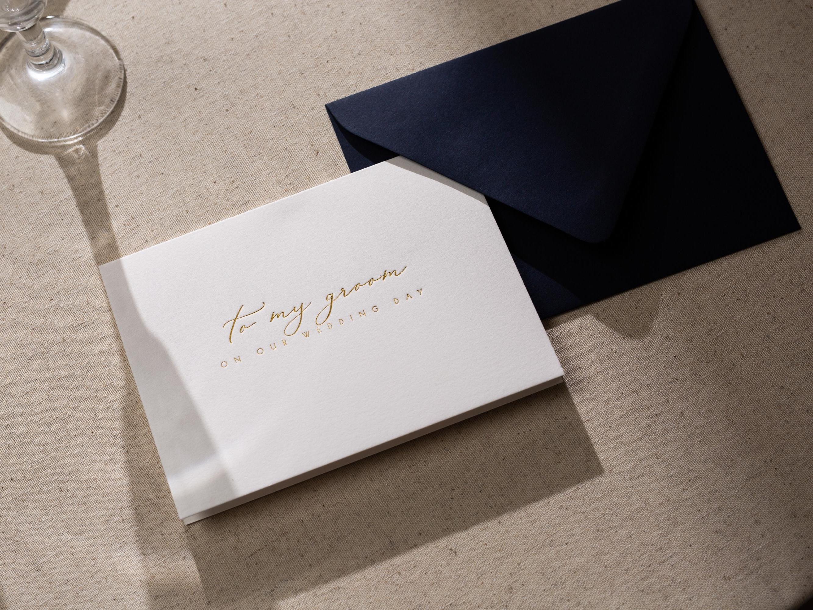May include: A white card with gold lettering that says "To my groom on our wedding day". The card is sitting on a beige surface with a dark blue envelope behind it.