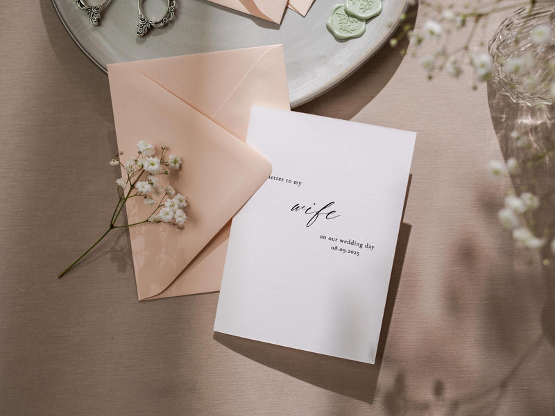 May include: A white card with black calligraphy that reads "Letter to my wife on our wedding day 08.09.2023". The card is on a light pink envelope and a light brown surface.