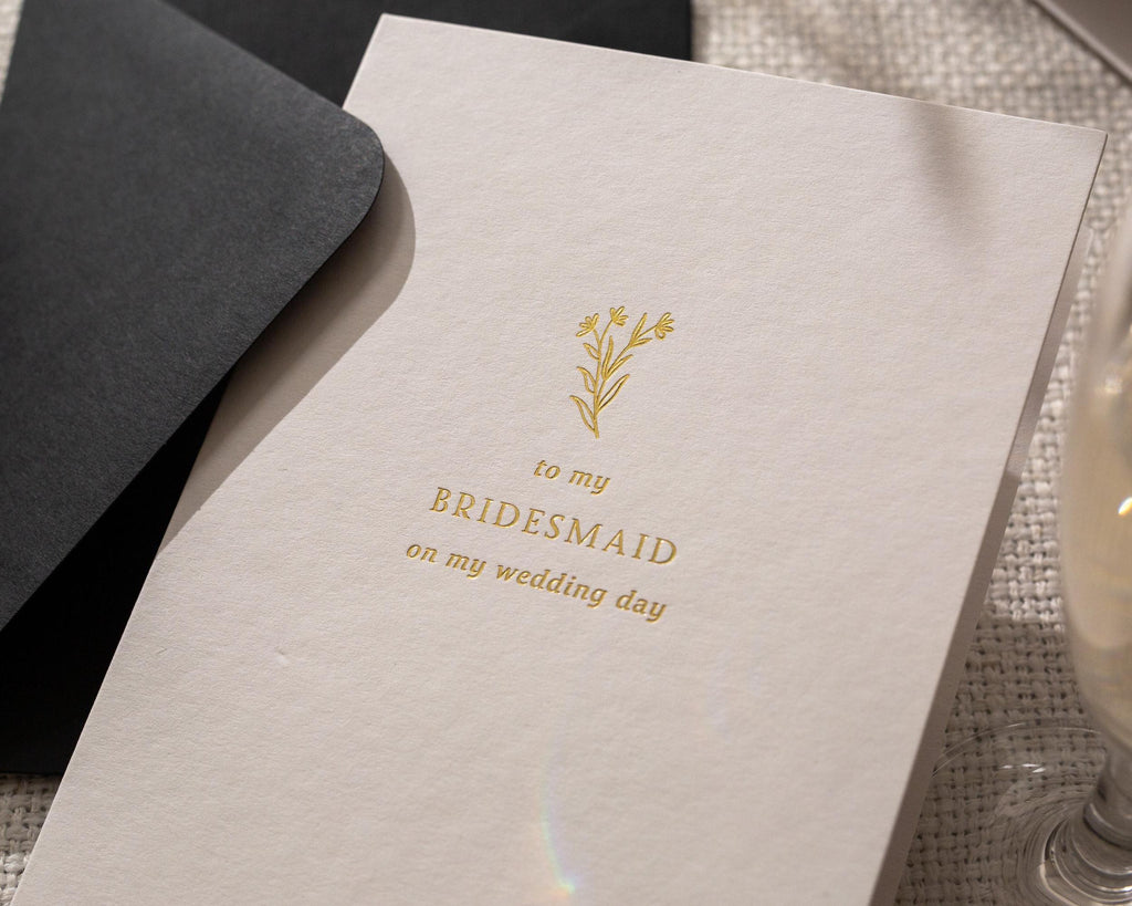 May include: A light beige card with gold text and a floral illustration. The text reads "to my BRIDESMAID on my wedding day." A black envelope is partially visible in the background.