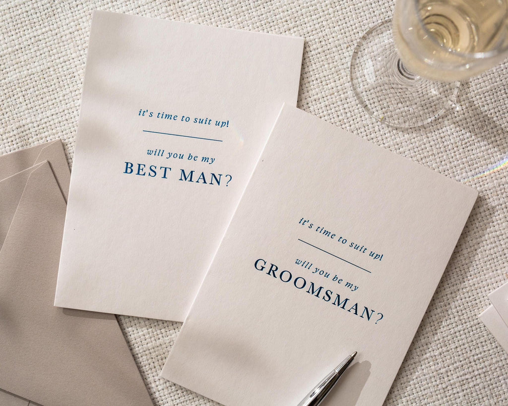 May include: Two white cards with blue text that read "It's time to suit up! Will you be my Best Man?" and "Will you be my Groomsman?" on a textured surface. A pen and a glass of champagne are also visible.