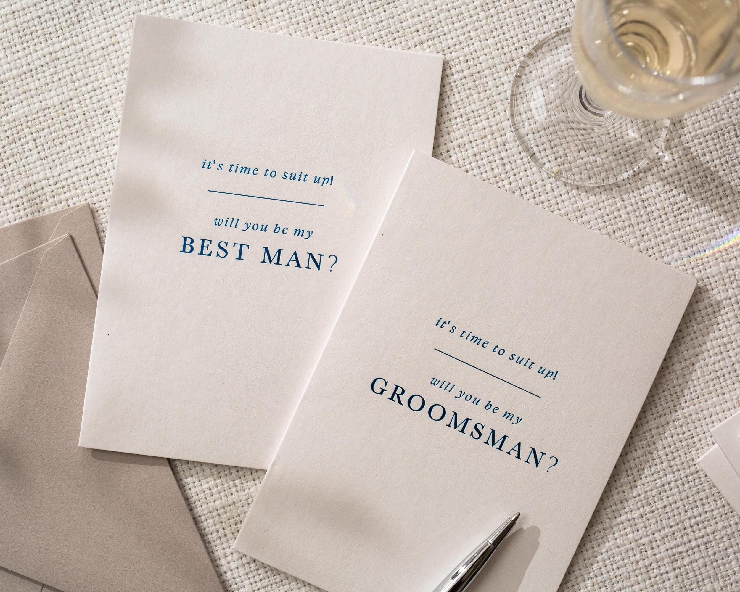May include: Two white cards with blue text that read "It's time to suit up! Will you be my Best Man?" and "Will you be my Groomsman?" on a textured surface. A pen and a glass of champagne are also visible.