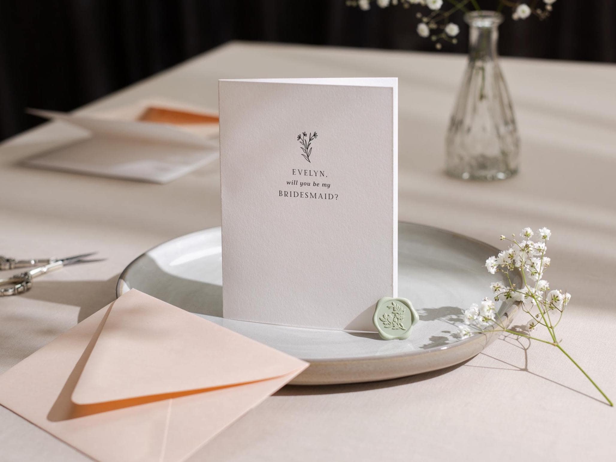 May include: A white card with a floral design and the text "EVELYN, will you be my BRIDESMAID?" on it. The card is on a light gray plate with a green wax seal.