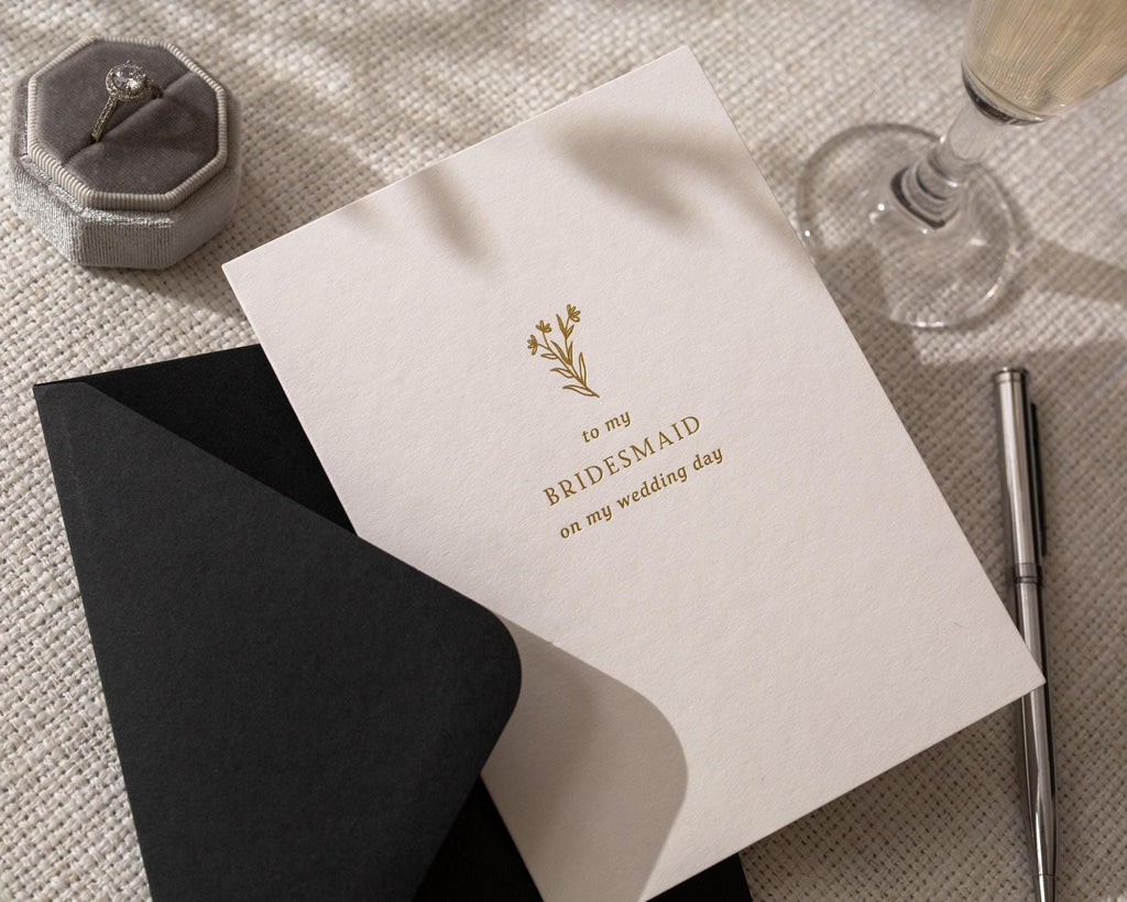 May include: A wedding card with the text "to my BRIDESMAID on my wedding day" in gold lettering, with a floral design. A black envelope is partially visible. A silver pen and a ring in a gray velvet box are also in the image.