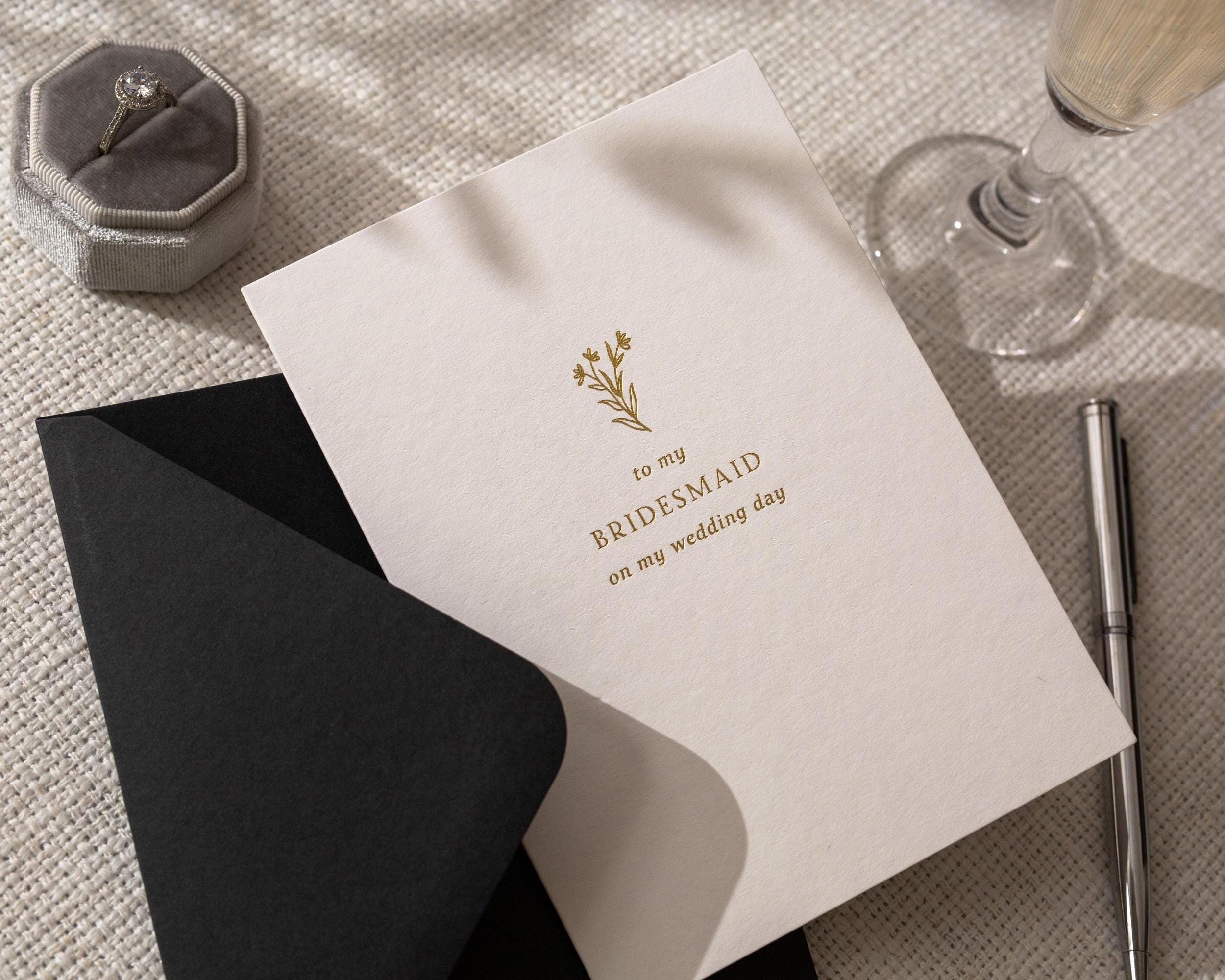 May include: A wedding card with the text "to my BRIDESMAID on my wedding day" in gold lettering, with a floral design. A black envelope is partially visible. A silver pen and a ring in a gray velvet box are also in the image.
