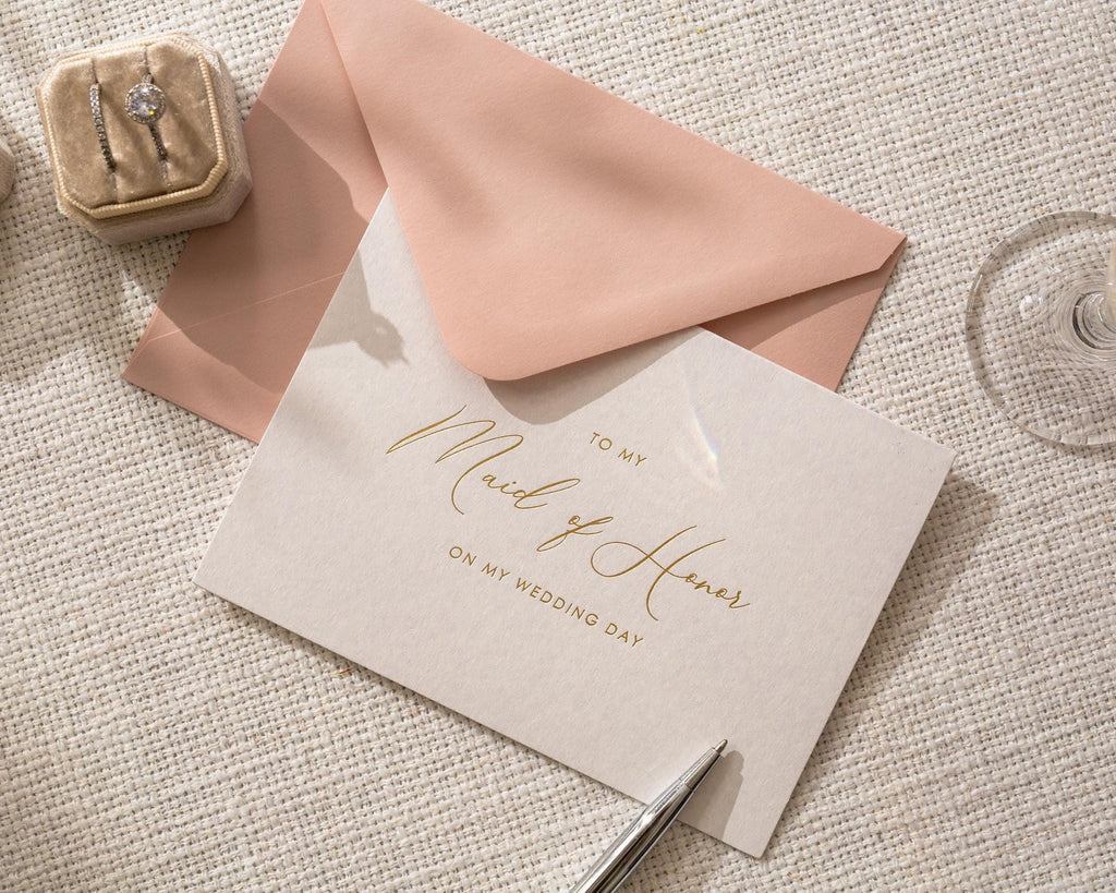 May include: A wedding invitation card with gold script that reads "To My Maid of Honor on My Wedding Day" is paired with a pink envelope. A small velvet ring box with two rings and a wine glass are also in the image.