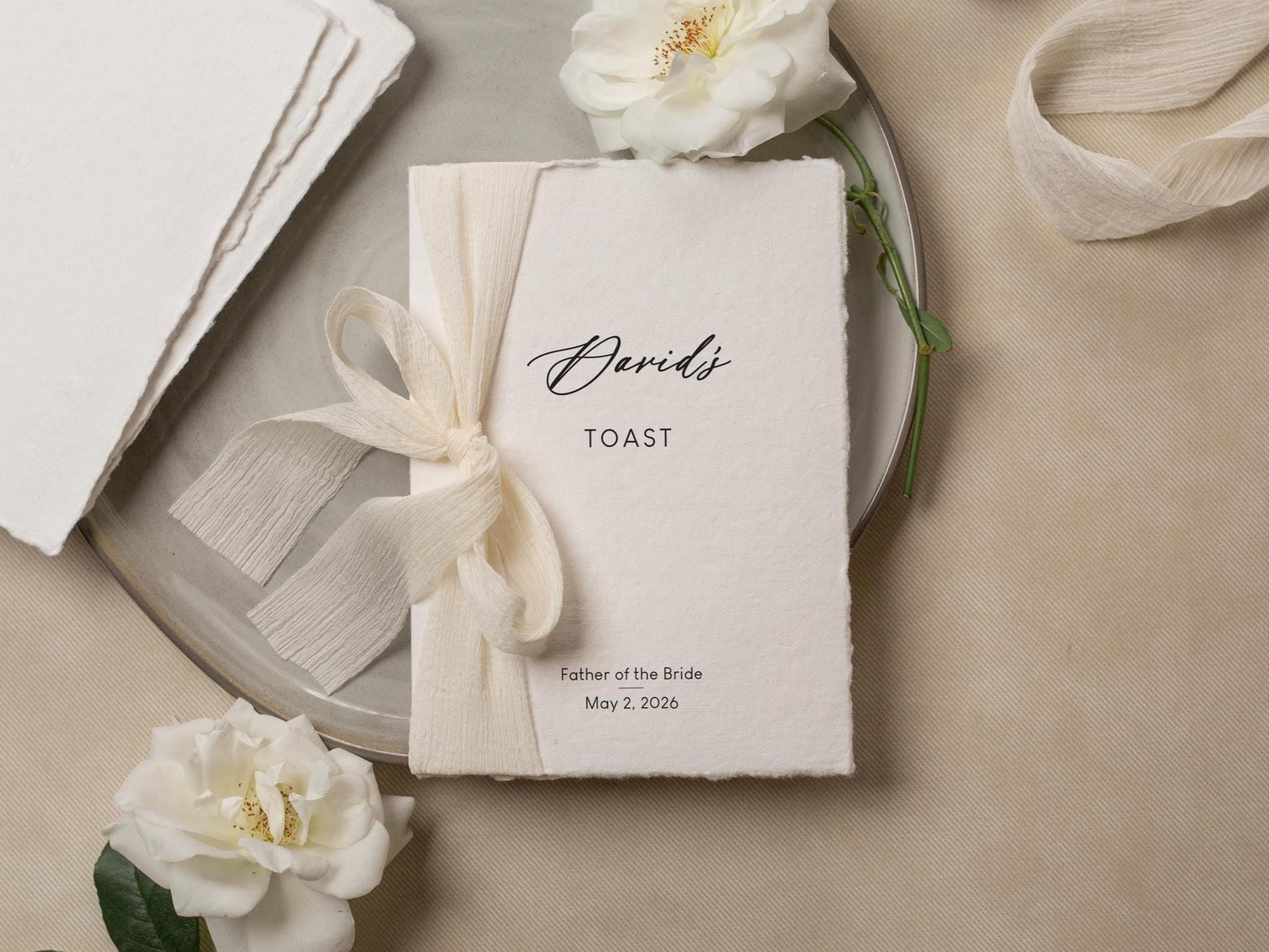 May include: A wedding toast card with the name "David's" in elegant script, tied with a cream ribbon. The card is on a plate with white roses. The text "TOAST" and "Father of the Bride May 2, 2026" are also visible.