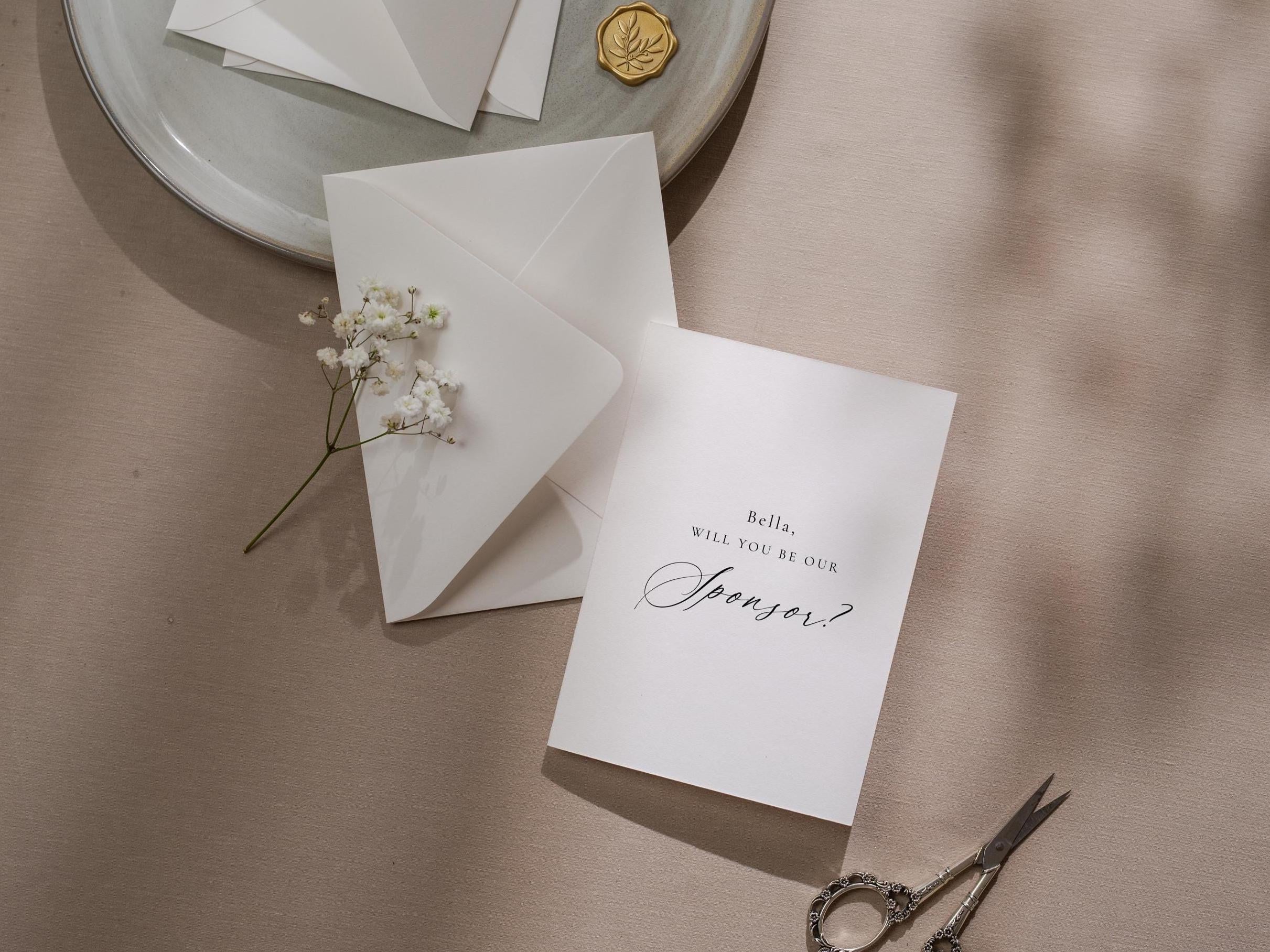 May include: A white invitation card with the text "Bella, WILL YOU BE OUR Sponsor?" is displayed next to a white envelope and a sprig of white flowers. A pair of ornate scissors and a gold wax seal are also visible.