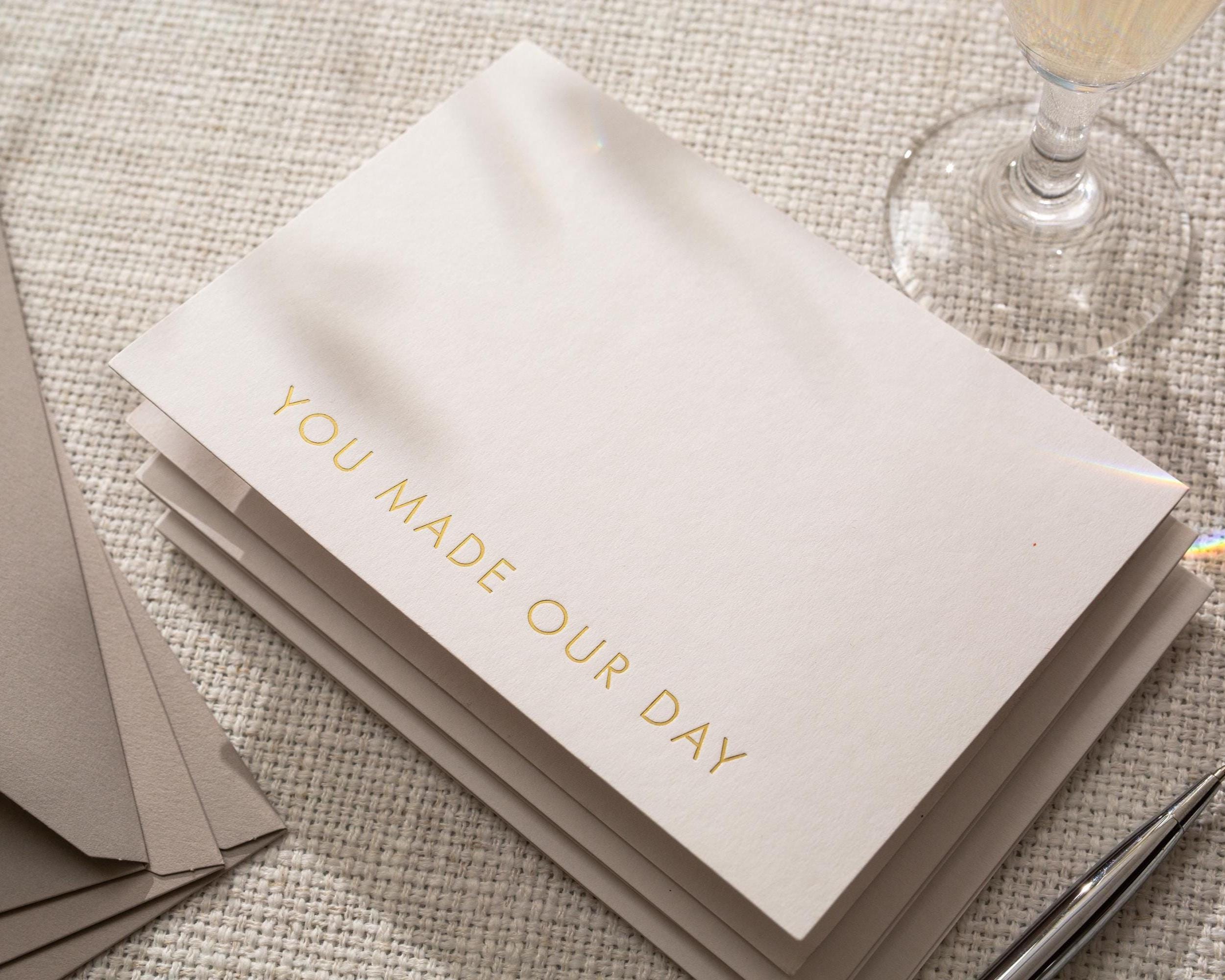 May include: A stack of ivory-colored cards with the gold-lettered phrase "YOU MADE OUR DAY." The cards are stacked on a textured surface, with a champagne glass and pen visible in the background. Additional gray envelopes are also present.
