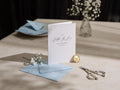 May include: A white card with the text "To my Father In Law On my Wedding Day 22.07.2023" in black calligraphy. The card is on a light blue envelope with a gold wax seal and a pair of silver scissors.