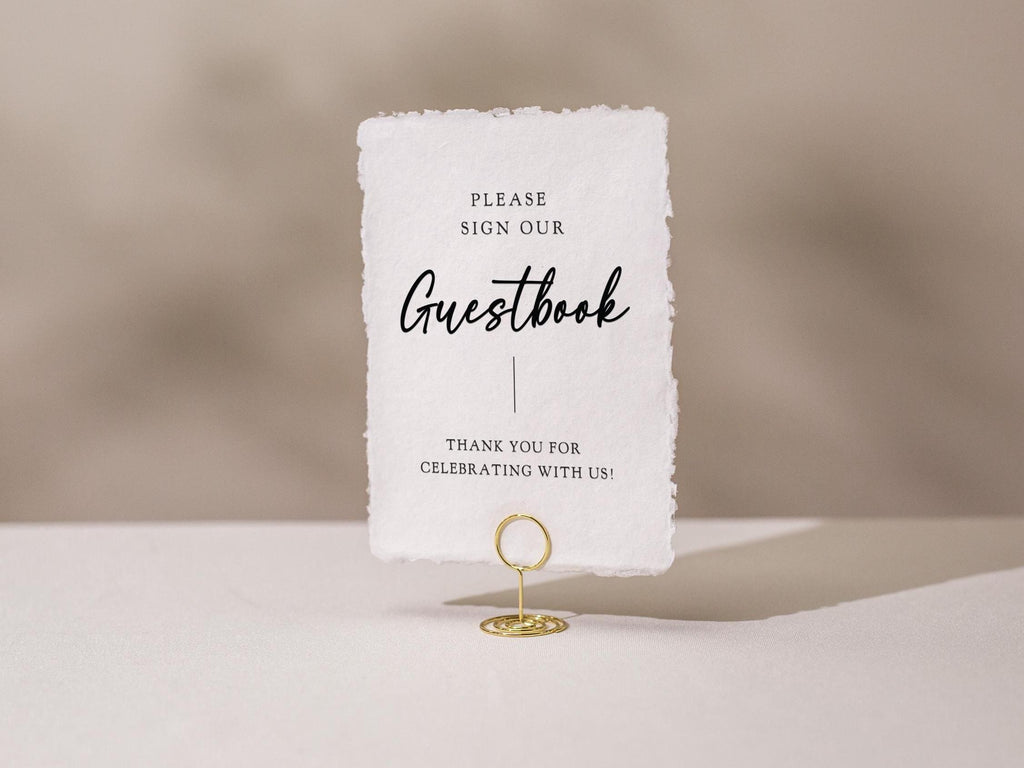 May include: A white rectangular sign with the words "PLEASE SIGN OUR Guestbook" in black script. The sign is held by a gold wire holder. The bottom of the sign reads "THANK YOU FOR CELEBRATING WITH US!"