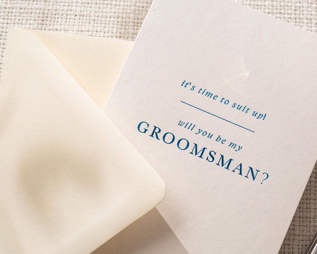 May include: A white card with blue text, and a cream-colored envelope. The card reads "it's time to suit up! will you be my GROOMSMAN?" The envelope is partially open, revealing the card.