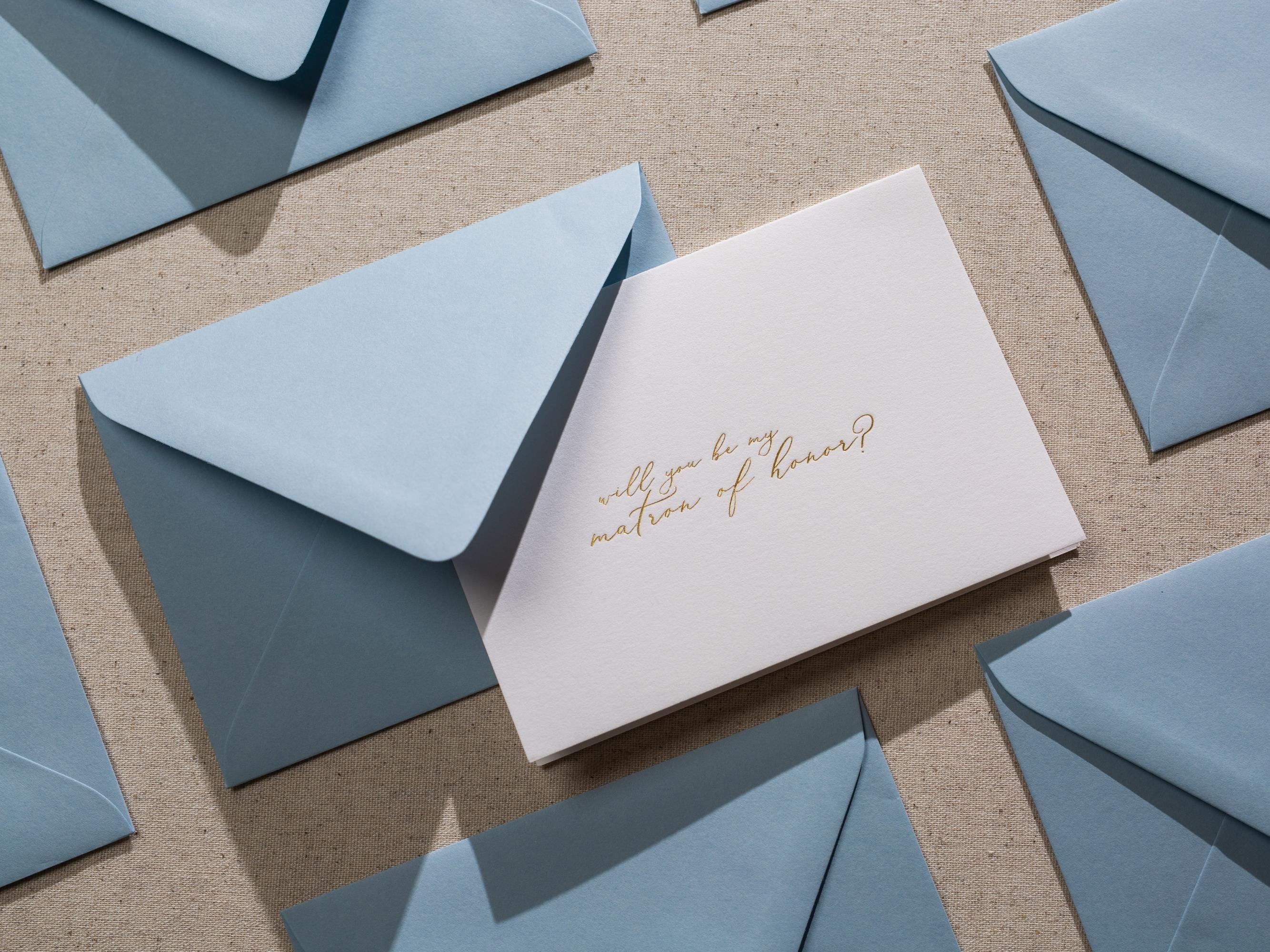 May include: A white card with gold lettering that says "Will you be my matron of honor?" The card is surrounded by light blue envelopes.