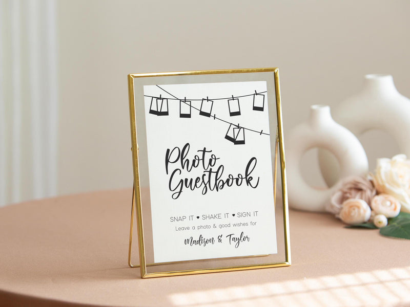 May include: A gold-framed sign with a white background and black text that reads "Photo Guestbook". The sign has a string of hanging photos with a black outline. The text below the photos reads "Snap it Shake it Sign it Leave a photo & good wishes for Madison & Taylor".
