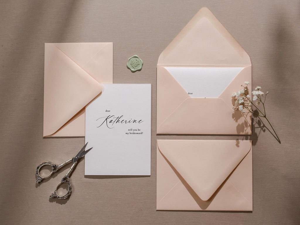 May include: A white card with black calligraphy that reads "Dear Katherine, will you be my bridesmaid?"  The card is surrounded by three pink envelopes and a small green wax seal.