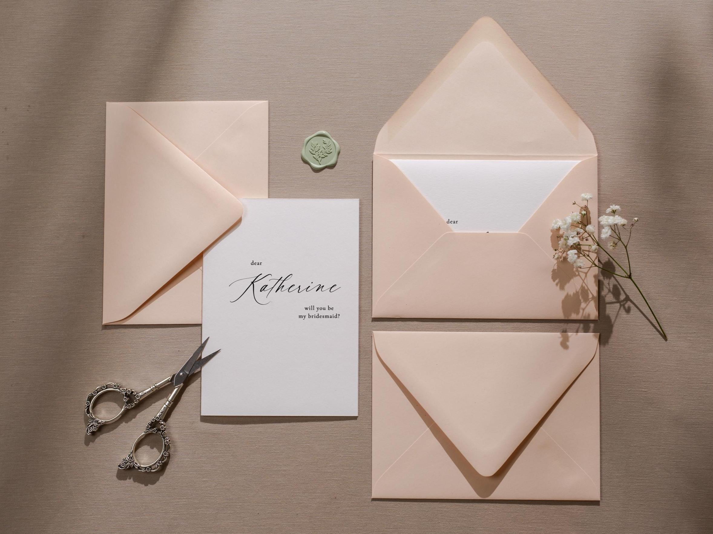 May include: A white card with black calligraphy that reads "Dear Katherine, will you be my bridesmaid?"  The card is surrounded by three pink envelopes and a small green wax seal.