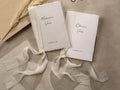 May include: Two white paper vow books with a cream colored ribbon tied around them. The books are labeled 'Madison's Vows' and 'Oliver's Vows' with the date 'November 22, 2023' printed below the names. The books are on a beige surface.