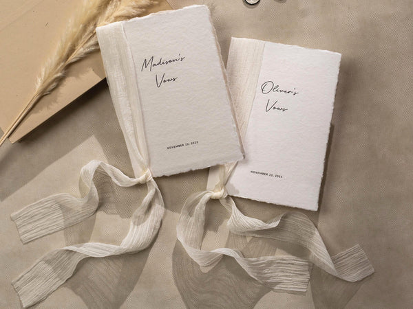 May include: Two white paper vow books with a cream colored ribbon tied around them. The books are labeled 'Madison's Vows' and 'Oliver's Vows' with the date 'November 22, 2023' printed below the names. The books are on a beige surface.
