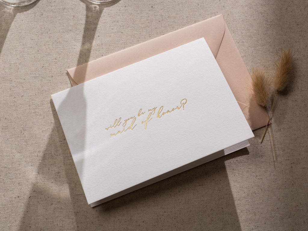 May include: White card with gold lettering that says "Will you be my maid of honor?"