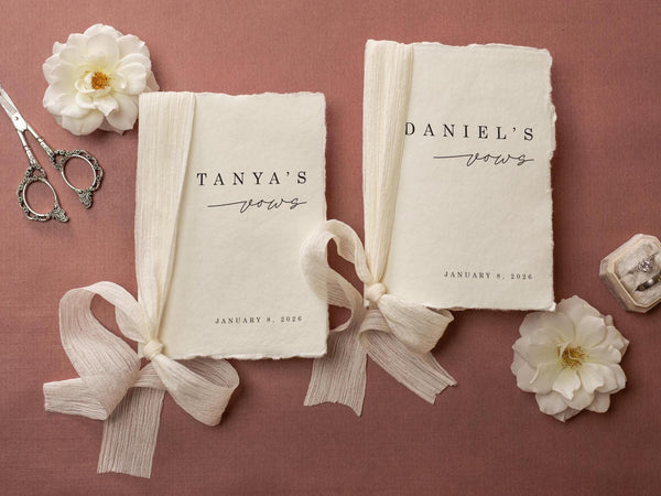 May include: Two cream-colored vow books with the names "Tanya's" and "Daniel's" printed in black script. Each book is tied with a cream ribbon. The date "January 8, 2026" is printed below the vows. The background is a muted rose color.