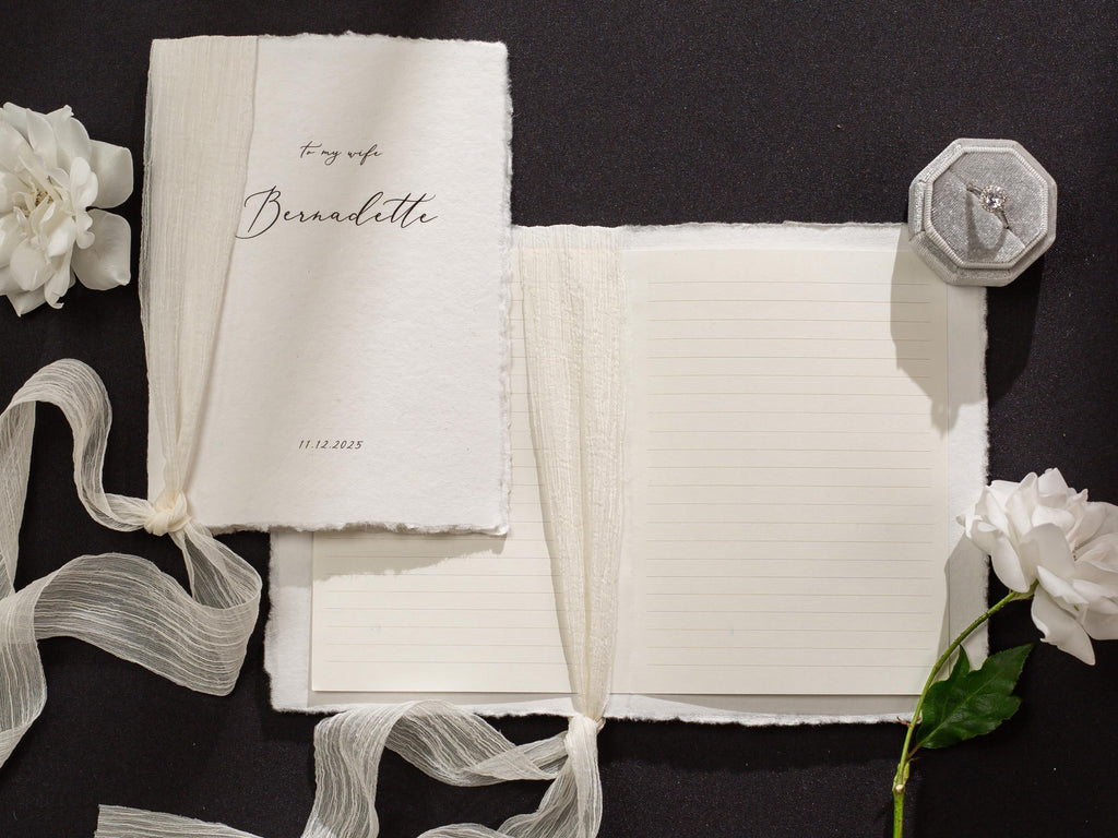 May include: A white journal with a ribbon closure and a handwritten inscription that reads "To my wife Bernadette 11.13.2023". The journal is open to reveal lined pages.