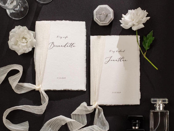 May include: Two white paper vow books with a white ribbon tied around them. The books have the text "to my wife Bernadette" and "to my husband Jonathan" written on them. The date "11.12.2023" is written on both books.