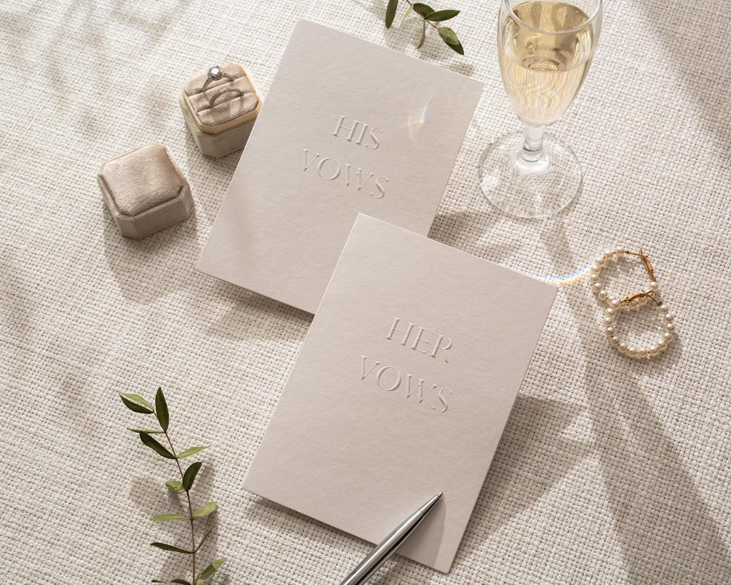 May include: A wedding scene with two vow books, one reading "HIS VOWS" and the other "HER VOWS". A ring box, a glass of champagne, and a pair of pearl earrings are also present. A pen rests near the vow books, and a sprig of greenery adds a natural touch.