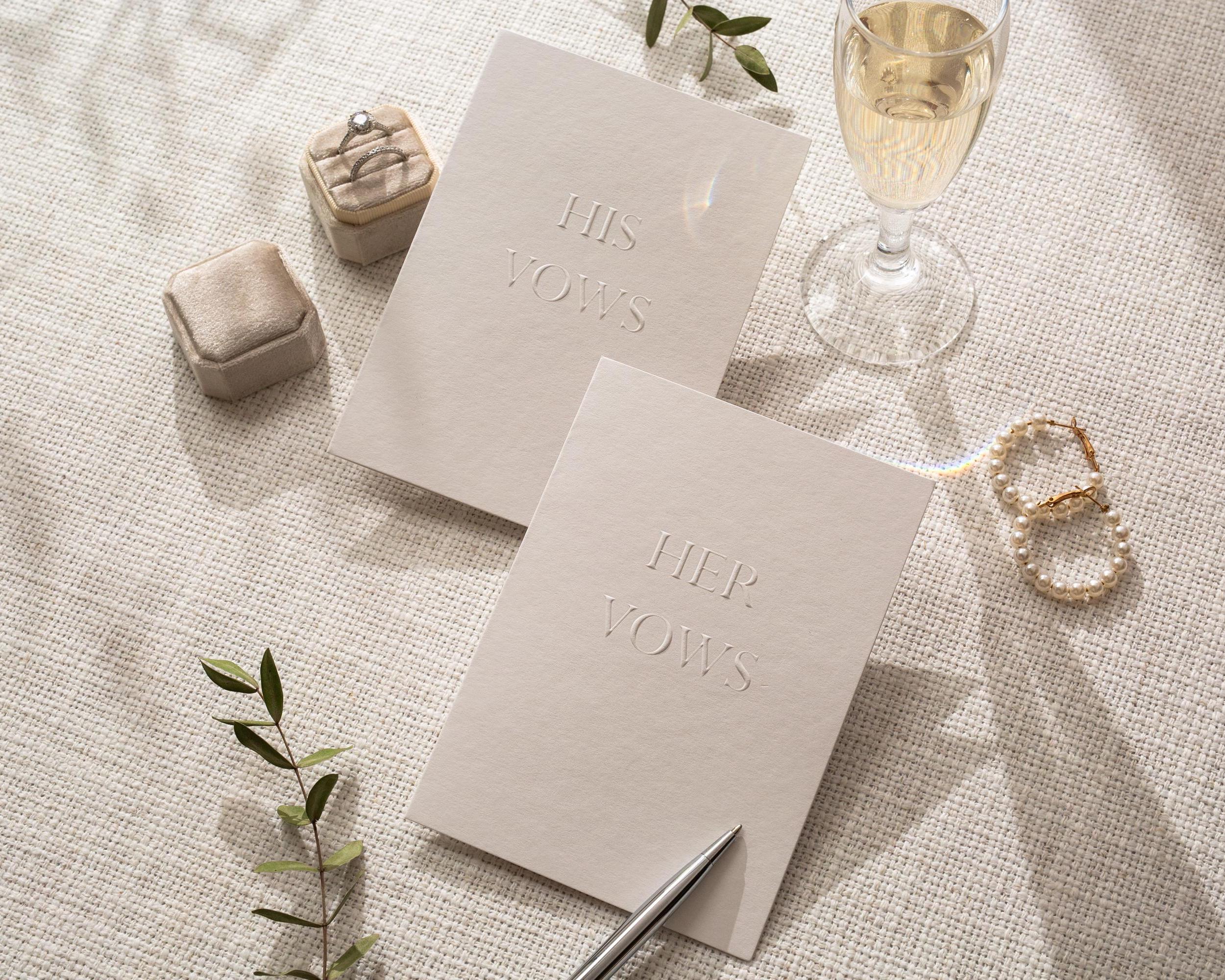 May include: A wedding scene with two vow books, one reading "HIS VOWS" and the other "HER VOWS". A ring box, a glass of champagne, and a pair of pearl earrings are also present. A pen rests near the vow books, and a sprig of greenery adds a natural touch.