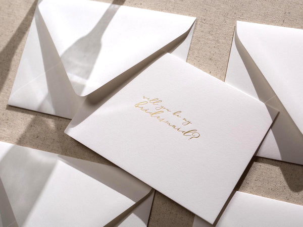 May include: A white card with gold foil lettering that says 'Will you be my bridesmaid?'
